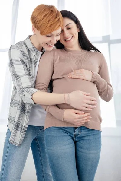 Mujer sonriendo mientras toca el vientre de la novia embarazada en casa - foto de stock