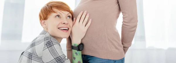 Panoramic shot of smiling woman touching tummy of pregnant girlfriend at home — Stock Photo