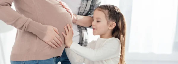 Panoramic shot of child and parent touching belly of pregnant woman in living room — Stock Photo