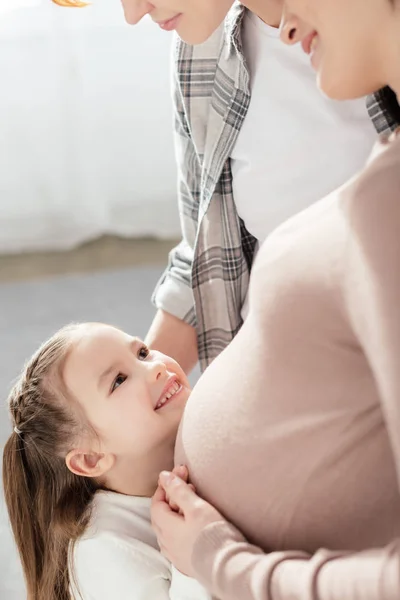 Вид сбоку на улыбающегося ребенка, трогающего живот беременной матери дома — стоковое фото