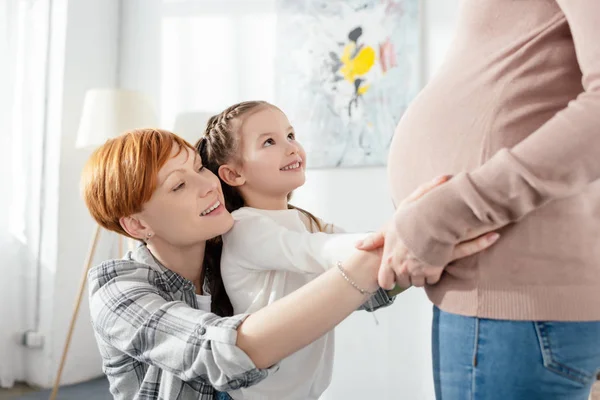 Vista lateral de la madre y la hija sonrientes tocando el vientre de la mujer embarazada en casa - foto de stock