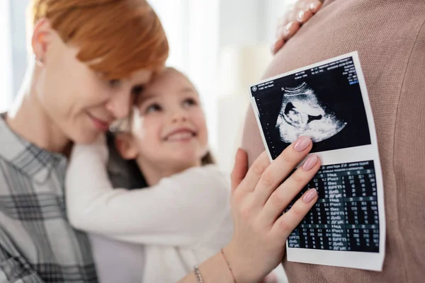 Селективное внимание матери, обнимающей улыбающуюся дочь и проводящей ультразвуковое сканирование ребенка возле живота беременной женщины — стоковое фото