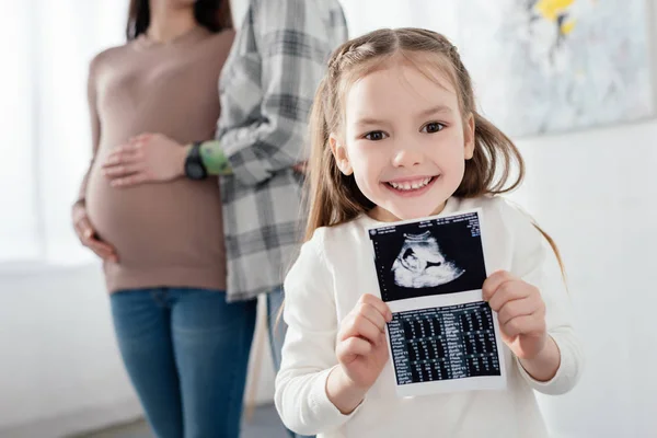 Selective focus of smiling kid holding ultrasound scan of baby near mom embracing pregnant woman at home — Stock Photo