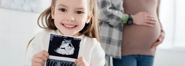Selective focus of smiling kid holding ultrasound scan of baby near mother embracing pregnant woman at home, panoramic shot — Stock Photo