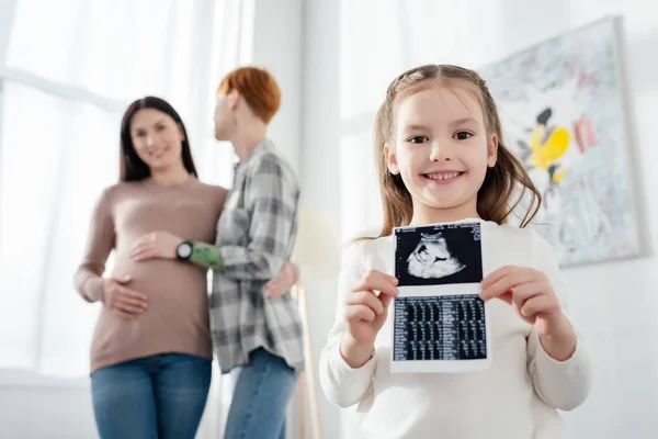 Выборочный фокус улыбающегося ребенка, держащего ультразвуковое сканирование ребенка рядом с женщиной, обнимающей беременную девушку — стоковое фото