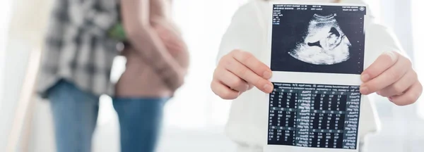 Cropped view of child holding ultrasound scan of baby while woman embracing pregnant girlfriend at home, panoramic shot — Stock Photo
