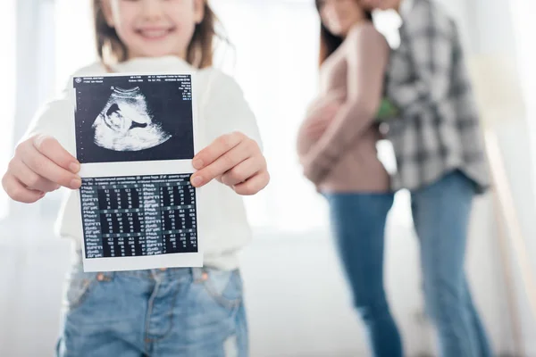Обрезанный вид улыбающегося ребенка, показывающего ультразвуковое сканирование ребенка, пока женщина обнимает беременную девушку дома — стоковое фото