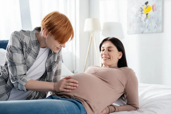 Smiling woman touching belly of pregnant girlfriend on bed — Stock Photo