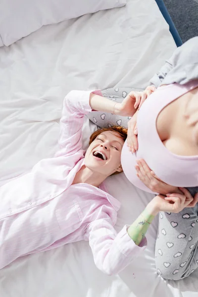 Top view of laughing woman in pajamas touching belly of pregnant girlfriend on bed — Stock Photo