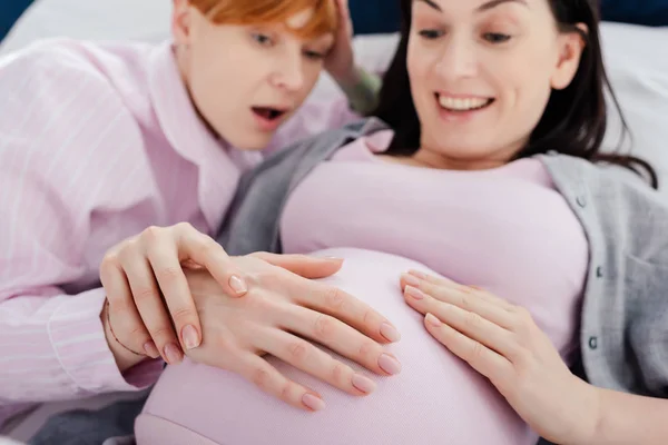 Focus sélectif de la femme choquée touchant le ventre de la petite amie enceinte souriante sur le lit sur fond noir — Photo de stock