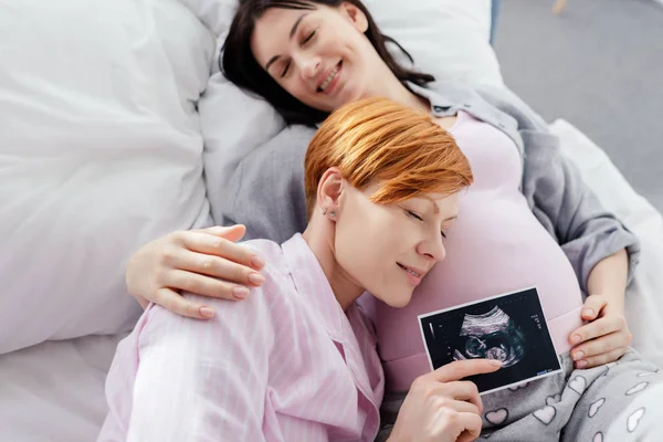 Smiling woman holding ultrasound scan of baby near belly of pregnant girlfriend on bed — Stock Photo