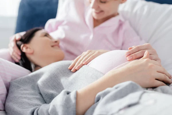 Selektiver Fokus einer lächelnden Frau, die ihre schwangere Freundin im Schlafanzug auf dem Bett ansieht — Stockfoto