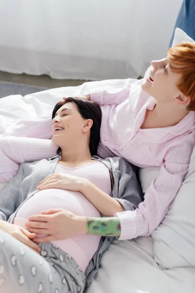 Lächelnde Frau berührt Bauch einer glücklichen schwangeren Freundin im heimischen Bett — Stockfoto