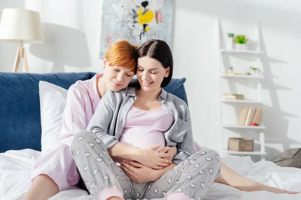 Lächelnde Frau umarmt schwangere Freundin im Bett — Stockfoto