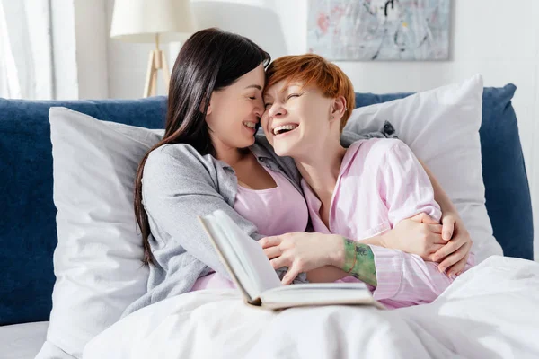Smiling woman embracing cheerful girlfriend near book on bed — Stock Photo
