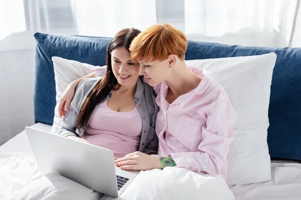Same sex couple in pajamas embracing and using laptop on bed — Stock Photo