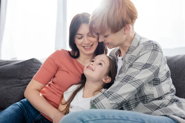 Same sex parents looking at smiling daughter on couch — Stock Photo