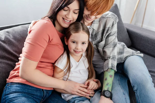Mães abraçando sorridente filha olhando para a câmera no sofá — Fotografia de Stock