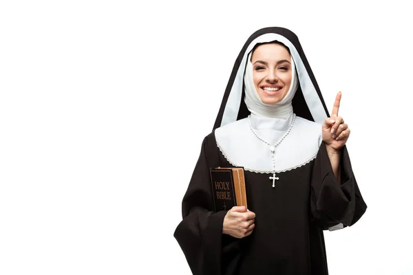 Monja alegre sosteniendo la biblia y apuntando hacia arriba con idea aislada en blanco - foto de stock