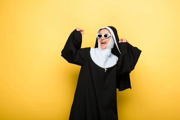 Attractive happy emotional nun dancing in sunglasses on yellow — Stock Photo