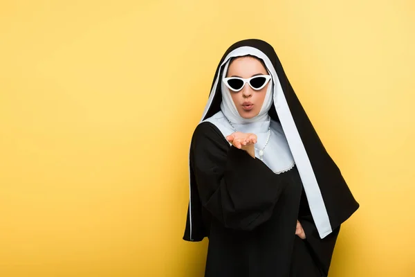 Monja atractiva en gafas de sol soplando aire beso en amarillo - foto de stock