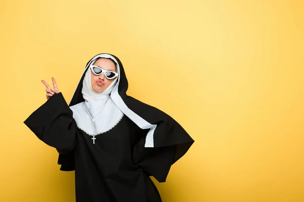 Attractive happy nun in sunglasses showing victory sign on yellow — Stock Photo