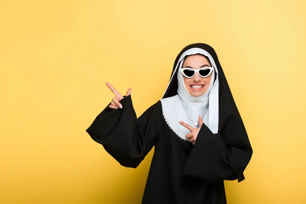 Привлекательная веселая монахиня в солнечных очках, указывающая на желтый цвет — стоковое фото
