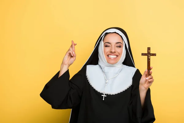 Joyeuse nonne tenant croix tout en montrant les doigts croisés sur jaune — Photo de stock