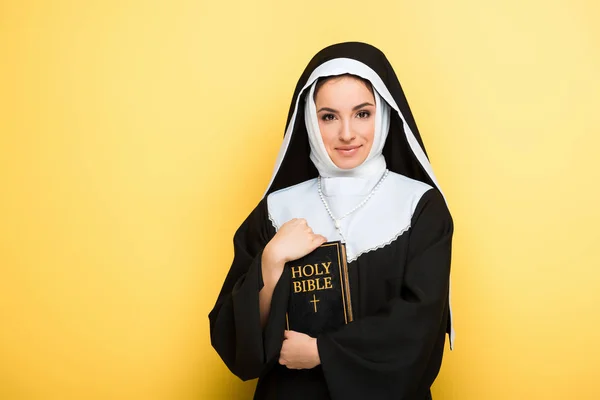 Attractive smiling nun holding holy bible on grey — Stock Photo