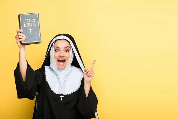 Atractiva monja excitada sosteniendo la sagrada biblia y señalando hacia arriba en gris - foto de stock
