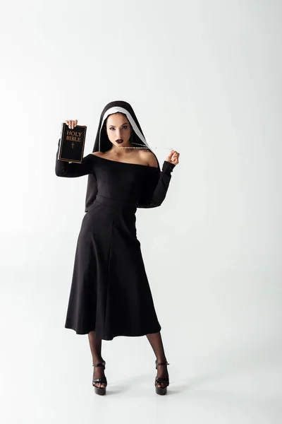 Hermosa monja sexy en vestido negro sosteniendo la biblia en gris - foto de stock