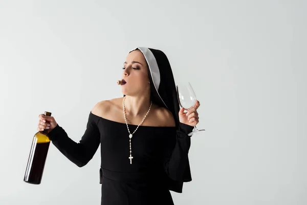 Attractive nun with cork in teeth holding glass and bottle of wine isolated on grey — Stock Photo