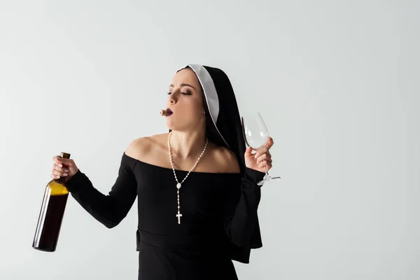 Jolie religieuse avec liège dans les dents tenant verre et bouteille de vin isolé sur gris — Photo de stock