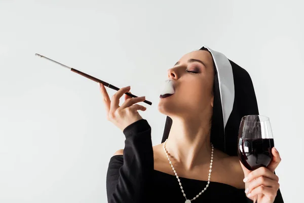 Attractive sexy nun with glass of wine smoking a cigarette in mouthpiece isolated on grey — Stock Photo