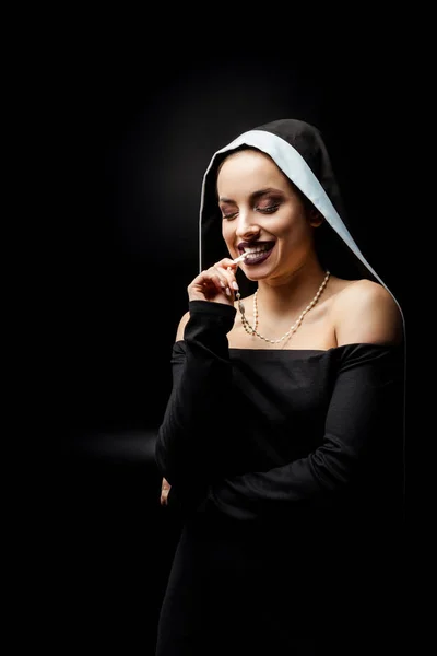 Сексуальна усміхнена черниця в чорній сукні кусає хрест, ізольована на чорному — стокове фото