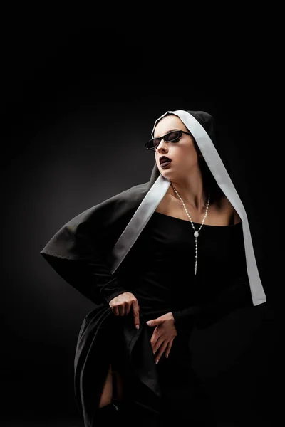 Sexy young nun in black dress and sunglasses, on black — Stock Photo
