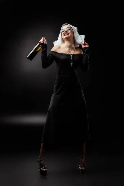 Excited sexy nun in sunglasses holding bottle of wine on grey — Stock Photo