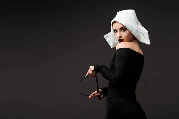 Sexy young nun in black dress holding sunglasses, isolated on grey — Stock Photo