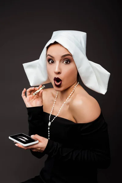 Шокированная монашка с банкнотой в долларах и смартфоном с кокаиновыми линиями на сером — стоковое фото