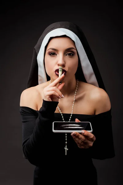 Emotional sexy nun holding dollar banknote and smartphone with cocaine lines showing silence symbol isolated on grey — Stock Photo