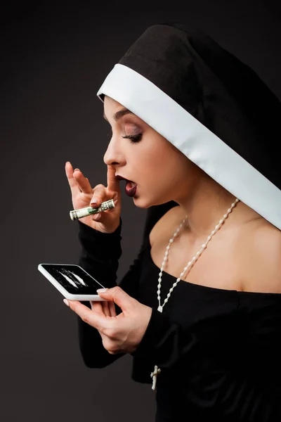 Sexy nun holding dollar banknote and smartphone with cocaine lines on grey — Stock Photo