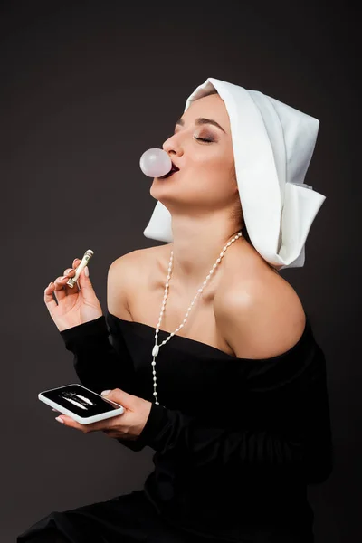 Sexy monja riendo con goma de mascar sosteniendo billete de dólar y teléfono inteligente con líneas de cocaína en gris - foto de stock