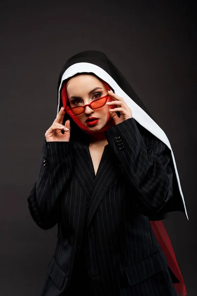 Modische sexy dämonische Nonne mit roten Augen posiert in schwarzem Anzug, roter Sonnenbrille und Schal, isoliert auf grau — Stockfoto