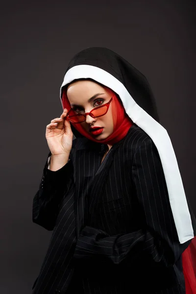 Modische sexy dämonische Nonne mit roten Augen posiert in schwarzem Anzug, roter Sonnenbrille und Schal, isoliert auf grau — Stockfoto