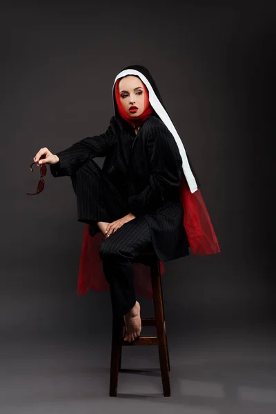 Pieds nus élégante nonne sexy posant en costume noir et écharpe rouge avec des lunettes de soleil, sur gris — Photo de stock