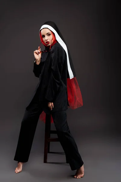Barefoot fashionable sexy nun posing in black suit and red scarf with sunglasses, on grey — Stock Photo