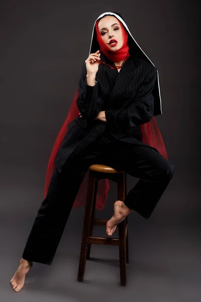 Barefoot fashionable sexy nun posing in black suit and red scarf with sunglasses, on grey — Stock Photo