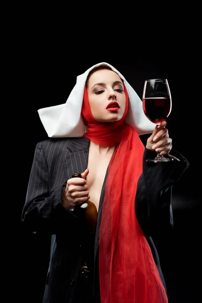 Monja sensual sosteniendo copa y botella de vino tinto aislado en negro - foto de stock