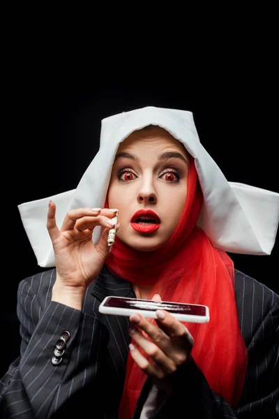 Shocked demonic nun holding dollar banknote and smartphone with cocaine lines isolated on black — Stock Photo