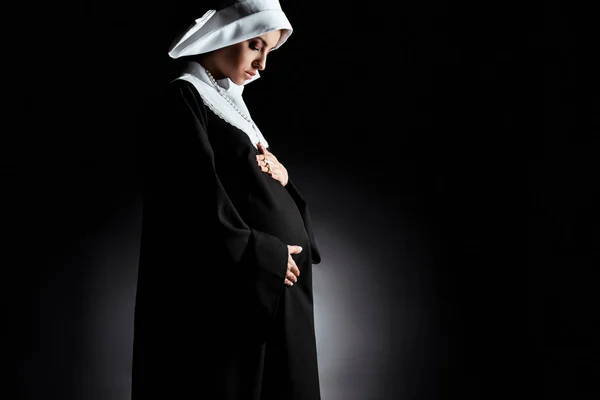 Attractive pregnant nun touching tummy on black — Stock Photo